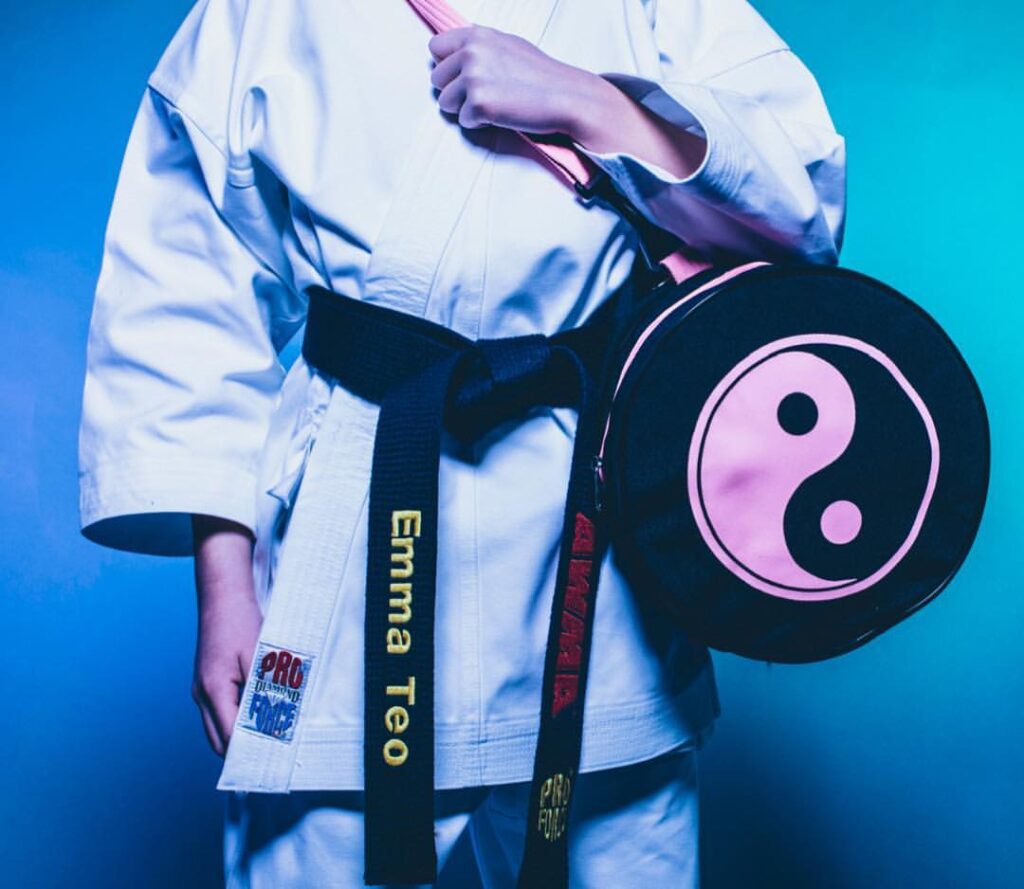 a torso wearing a white karate gi. the person is holding a black gear bag with a pink yin and yang printed on it. they're wearing a black belt that is embroidered with the AWMA logo and their name.